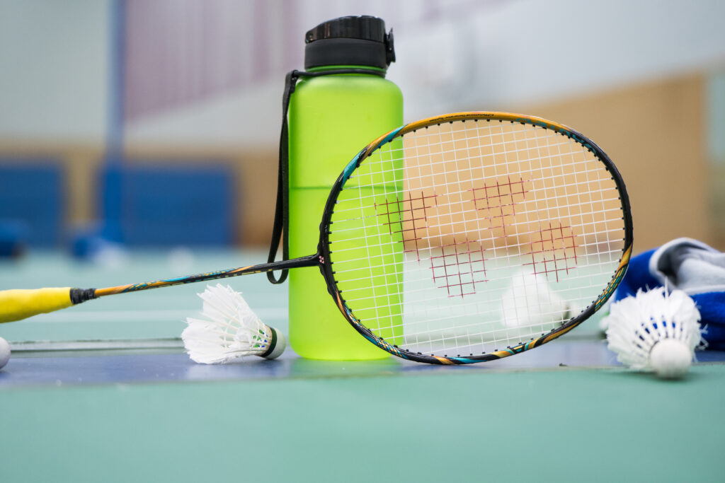 Badmintonschläger, Bälle und eine Flasche stehen in einer Halle auf dem Boden.